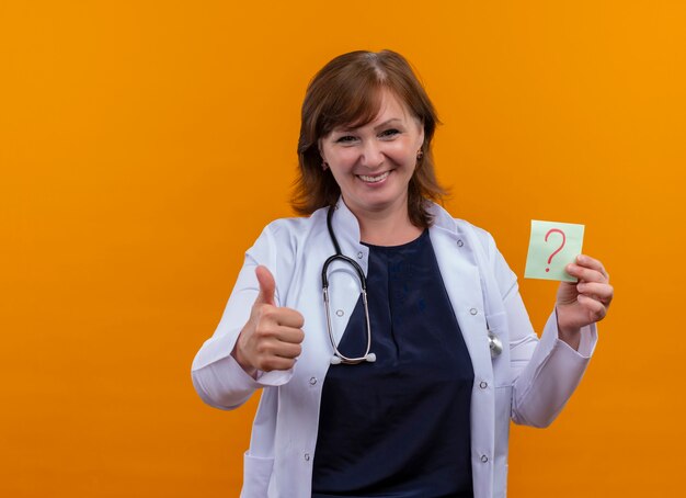 Sonriente mujer de mediana edad médico con bata médica y estetoscopio mostrando el pulgar hacia arriba y sosteniendo una nota de papel con signo de interrogación en la pared naranja aislada con espacio de copia