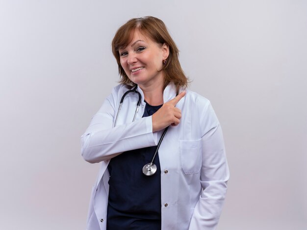 Sonriente mujer de mediana edad médico vistiendo bata médica y estetoscopio apuntando con el dedo al lado derecho en la pared blanca aislada con espacio de copia