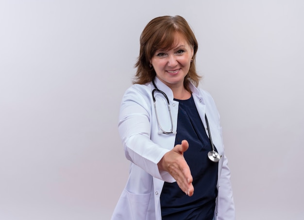 Sonriente mujer de mediana edad médico vistiendo bata médica y estetoscopio haciendo gesto de apretón de manos en la pared blanca aislada con espacio de copia