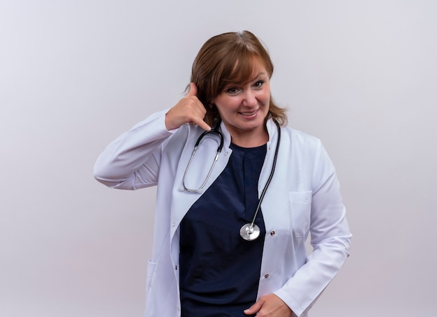Sonriente mujer de mediana edad médico vistiendo bata médica y estetoscopio haciendo gesto de teléfono en pared blanca aislada