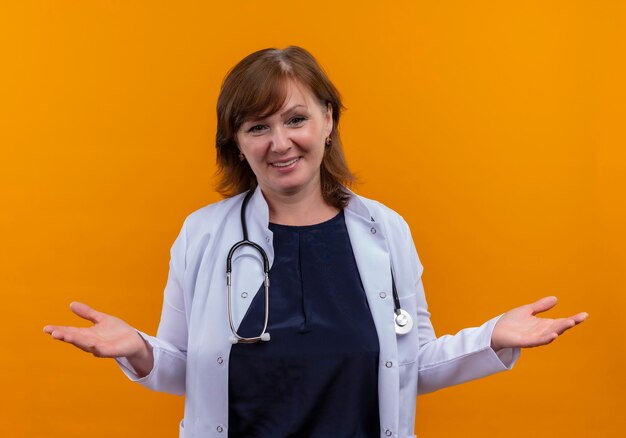 Sonriente mujer de mediana edad médico vistiendo bata médica y estetoscopio mostrando las manos vacías en la pared naranja aislada