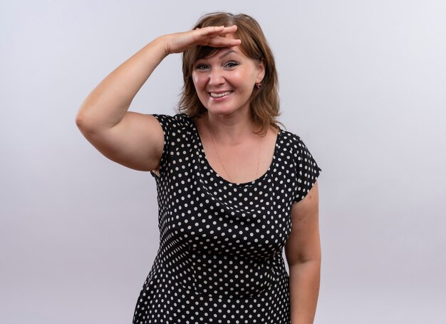 Sonriente mujer de mediana edad poniendo la mano cerca de la frente en la pared blanca aislada