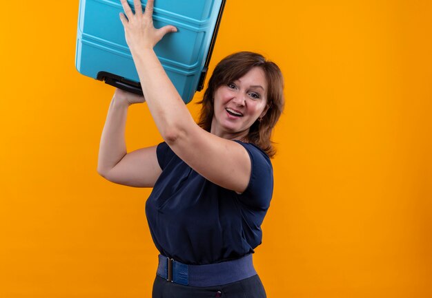 Sonriente mujer de mediana edad que viaja levantando la maleta en naranja aislada con espacio de copia