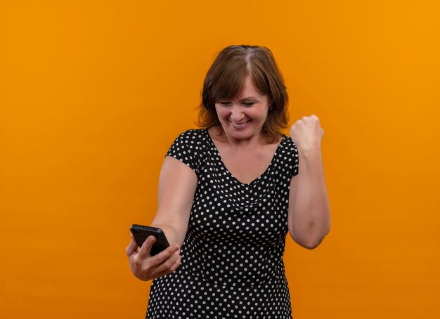 Sonriente mujer de mediana edad sosteniendo el teléfono móvil con el puño levantado en la pared naranja aislada con espacio de copia