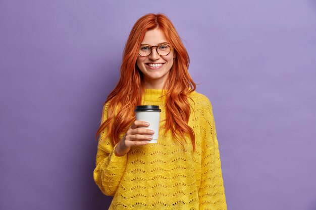 Sonriente mujer milenaria pelirroja sostiene una taza de café y tiene buen humor, disfruta de la pausa para el almuerzo, expresa emociones positivas, visita, el mejor café para llevar, viste ropa casual.