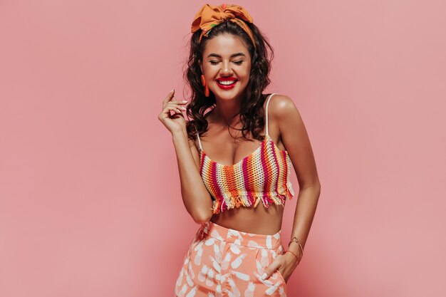 Sonriente mujer de moda con diadema naranja y lápiz labial rojo en top moderno a rayas y pantalones impresos posando con los ojos cerrados.