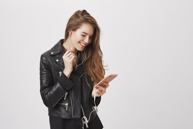 Sonriente mujer moderna con chaqueta de cuero, usa el teléfono móvil y escucha música en los auriculares