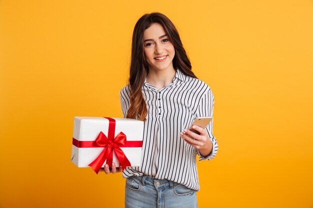 Sonriente mujer morena en camisa con caja de regalo y teléfono inteligente