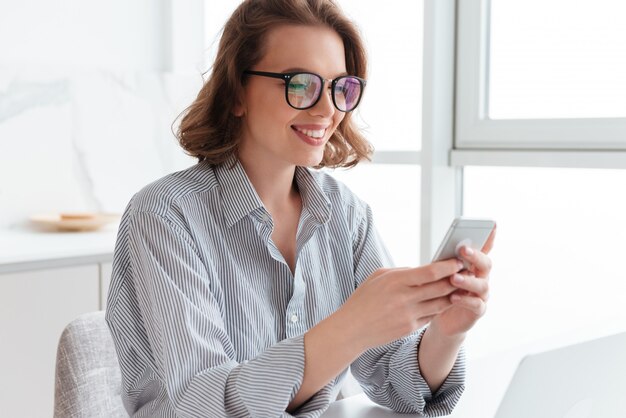 sonriente mujer morena con gafas mensaje de texto en el teléfono inteligente mientras está sentado en la cocina