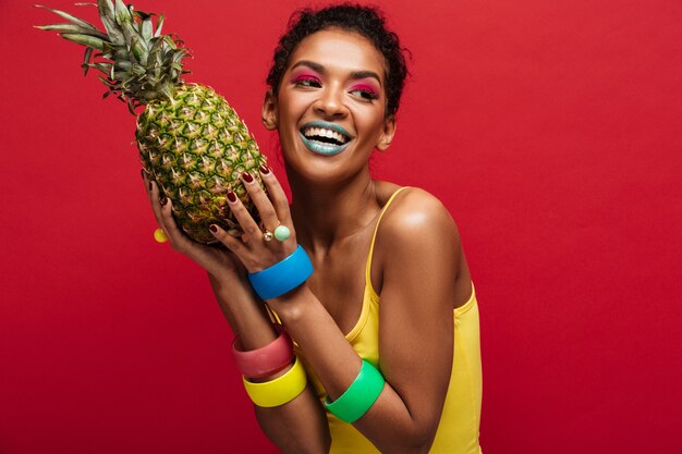 Sonriente mujer mulata con maquillaje de moda en camisa amarilla disfrutando de vitaminas naturales sosteniendo en manos piña madura fresca aislada, sobre pared roja