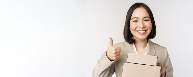 Sonriente mujer de negocios asiática mostrando los pulgares hacia arriba y cajas con productos de entrega preparan el pedido para el cliente de pie sobre fondo blanco.