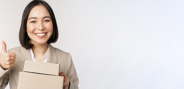 Sonriente mujer de negocios asiática mostrando los pulgares hacia arriba y cajas con productos de entrega preparan el pedido para el cliente de pie sobre fondo blanco.