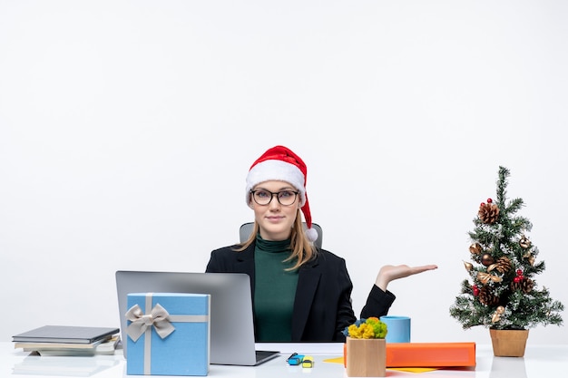 Sonriente mujer de negocios con un sombrero de santa claus sentado en una mesa con un árbol de Navidad y un regalo y apuntando algo en el lado izquierdo de la oficina sobre fondo blanco.