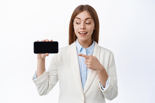 Sonriente mujer de negocios con traje profesional, apunta al teléfono inteligente vacío, sostiene la pantalla horizontal, introduce webiste o promoción en línea, pared blanca