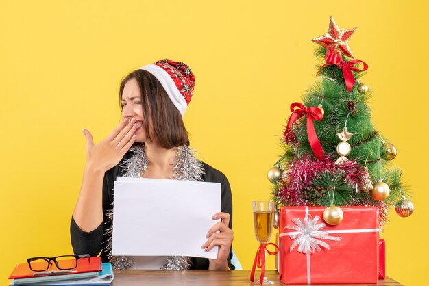 Sonriente mujer de negocios en traje con sombrero de santa claus y adornos de año nuevo trabajando solo sosteniendo documentos bostezando y sentado en una mesa con un árbol de Navidad en la oficina