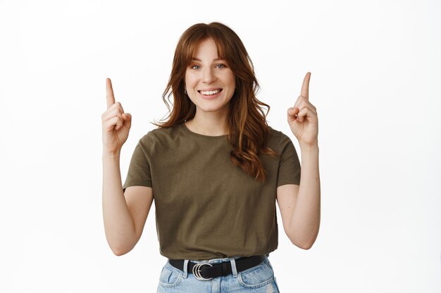 Sonriente mujer pelirroja apuntando con el dedo hacia arriba feliz, de pie en camiseta verde sobre blanco.