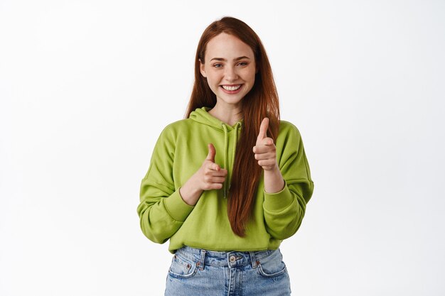 Sonriente mujer pelirroja señalando con el dedo, felicitándote, invitándote o eligiéndote, reclutando personas en blanco. Copia espacio