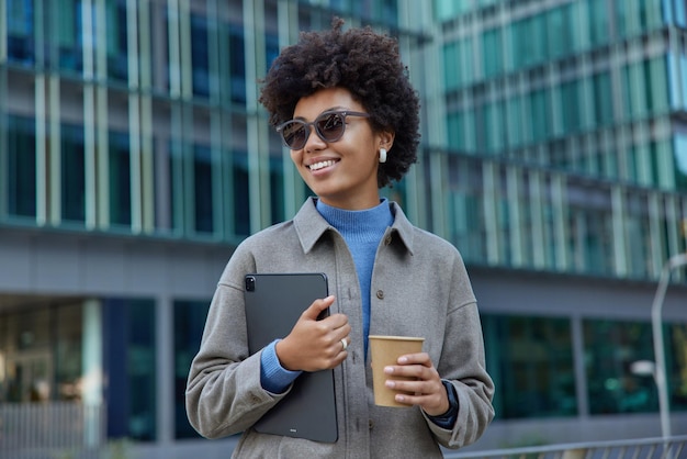 Sonriente mujer de pelo rizado sostiene una taza digital y de papel con café vestida con ropa de moda gafas de sol posa en el centro de la ciudad contra los rascacielos modernos que regresan del trabajo Concepto de estilo de vida