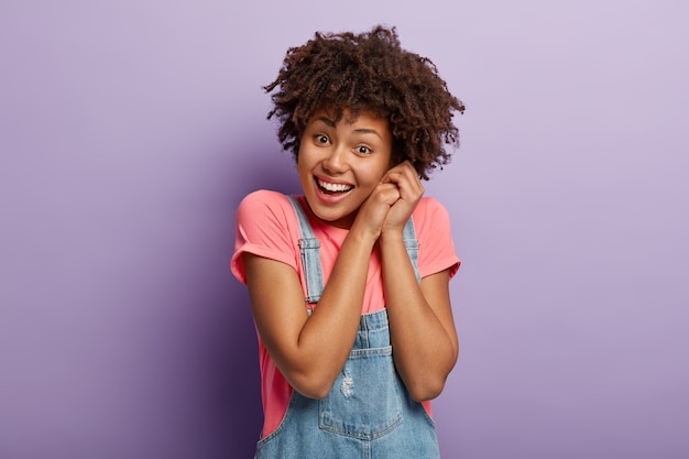 Sonriente mujer de piel oscura amigable mantiene las manos juntas suavemente cerca de la cabeza, mira a la cámara con satisfacción