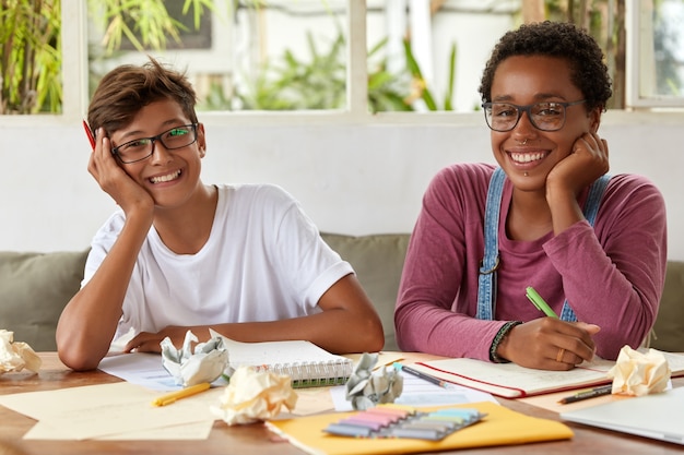 Sonriente mujer de piel oscura da buenos consejos a un compañero de clase masculino, habla sobre la tarea común, escribe registros en un cuaderno de espiral, habla sobre un proyecto común y hacen investigaciones o planes juntos