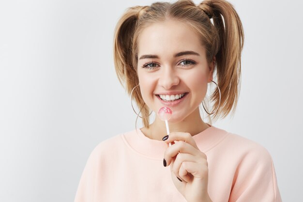 Sonriente mujer positiva con aspecto atractivo, vistiendo sudadera rosa suelta. Adolescente feliz con dos coletas en el pelo que muestra emociones positivas comiendo piruleta.