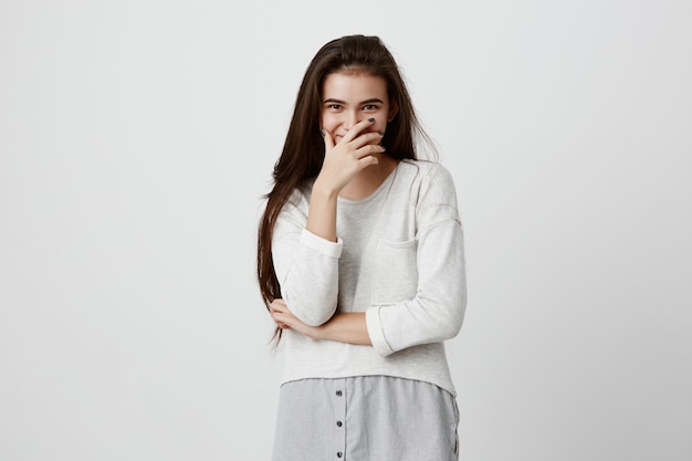 Sonriente mujer positiva con cabello largo oscuro, vistiendo suéter, posando contra la pared gris. Feliz mujer morena escondiendo la cara detrás de la mano, mostrando emociones positivas después de recibir un cumplido agradable