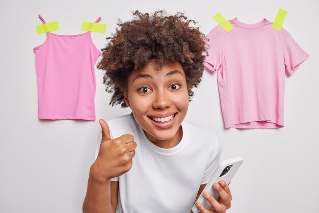 Sonriente mujer rizada positiva mantiene el pulgar hacia arriba recomienda que algo use celular moderno para vender ropa en línea en poses de tiendas de Internet contra fondo blanco con ropa colgada detrás