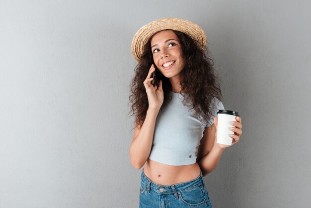 Sonriente mujer rizada con sombrero hablando por el teléfono inteligente con café en mano y mirando hacia arriba sobre fondo gris