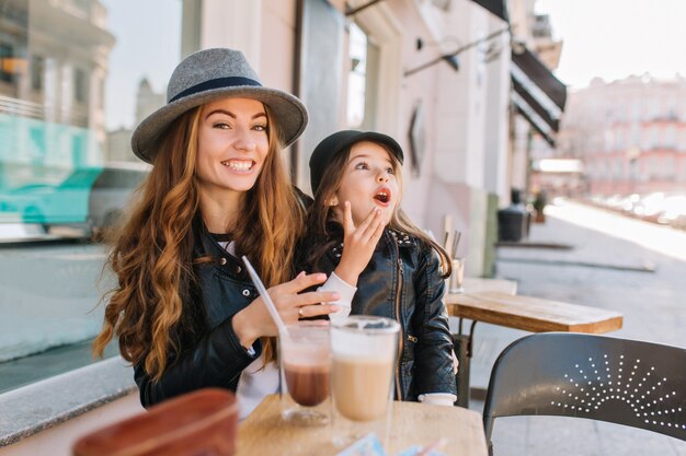 Sonriente mujer rizada con sombrero vintage y chaqueta de cuero posando con hija emocionada en la cafetería, mientras bebe café.