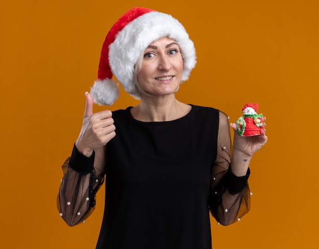 Sonriente mujer rubia de mediana edad con sombrero de navidad mirando a cámara mostrando una pequeña estatua de muñeco de nieve a la cámara y mostrando el pulgar hacia arriba aislado sobre fondo naranja