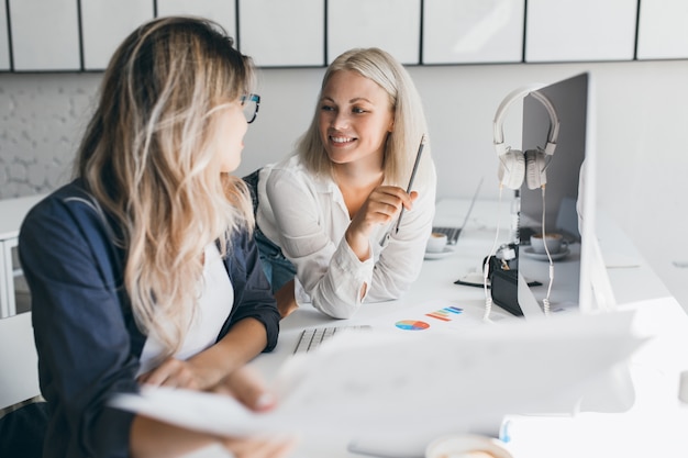 Sonriente mujer rubia de pelo corto hablando con su colega en la oficina mientras jugaba con lápiz. Retrato de interior de mujer diseñadora web mirando a mujer alegre con camisa blanca.