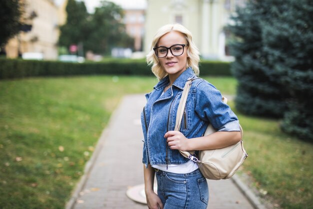 Sonriente mujer rubia en retrato de gafas en la ciudad vistiendo jeans suite en la mañana