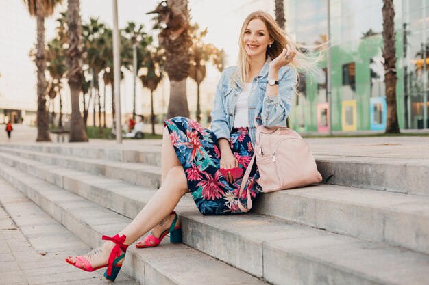 Sonriente mujer rubia sentada en las escaleras en las calles de la ciudad con elegante falda estampada y chaqueta vaquera de gran tamaño con mochila de cuero