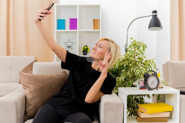 Sonriente mujer rusa rubia hermosa se sienta en el sillón gesticulando signo de mano de victoria mirando el teléfono