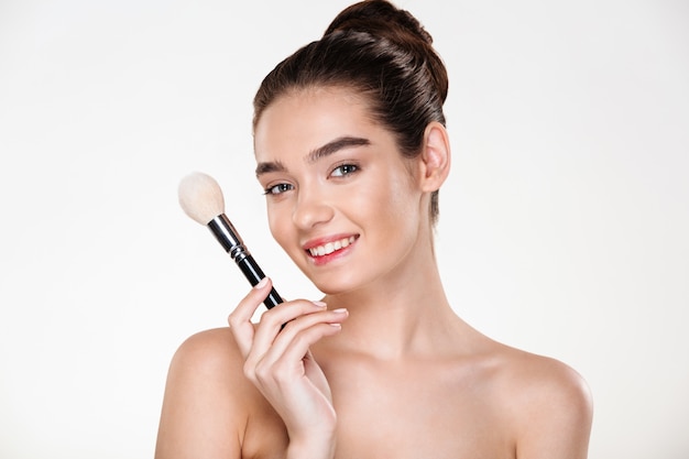 sonriente mujer semidesnuda con piel fresca con pincel para maquillaje cerca de la cara aplicando corrector