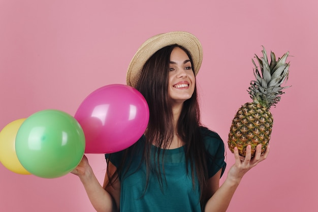 Sonriente mujer en un sombrero posa con globos de colores y una piña