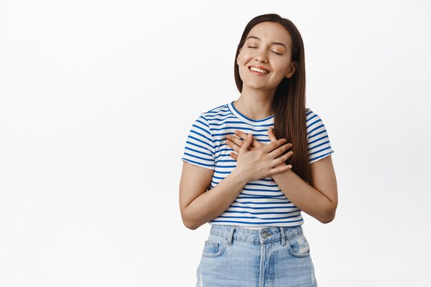 Sonriente mujer soñadora pensando en algo conmovedor, cogidos de la mano en el pecho del corazón, cerrar los ojos y soñar despierto, imaginando algo hermoso, pared blanca.