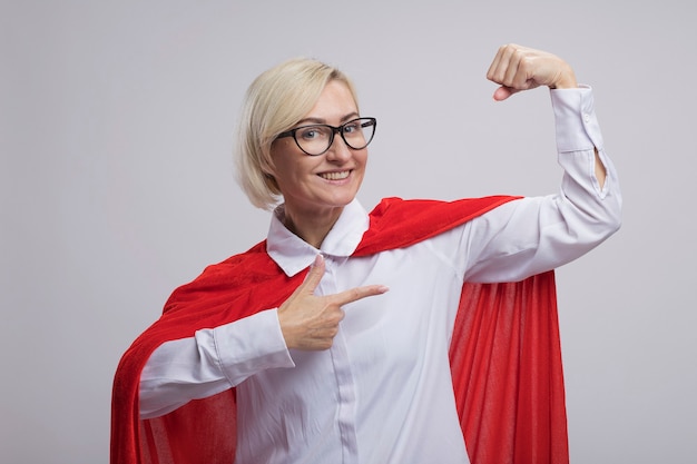 Sonriente mujer de superhéroe rubia de mediana edad en capa roja con gafas haciendo un gesto fuerte apuntando a sus músculos aislados en la pared blanca