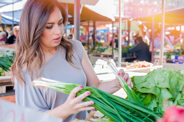 Sonriente mujer con vegetales en la tienda del mercado. Mujer que elige las verduras frescas en el mercado verde. Retrato de mujer joven hermosa que elige verduras de hoja verde