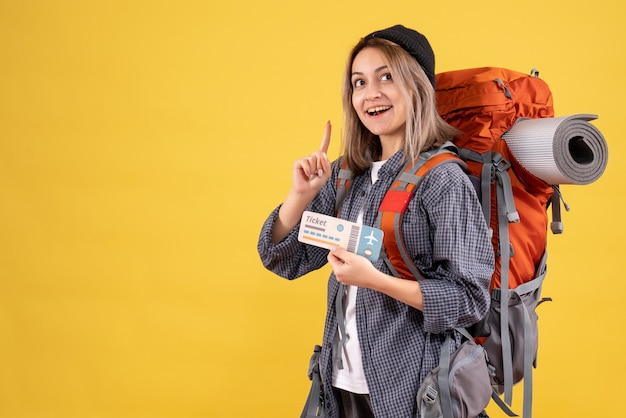 sonriente mujer viajera con mochila sosteniendo el billete apuntando con el dedo hacia arriba