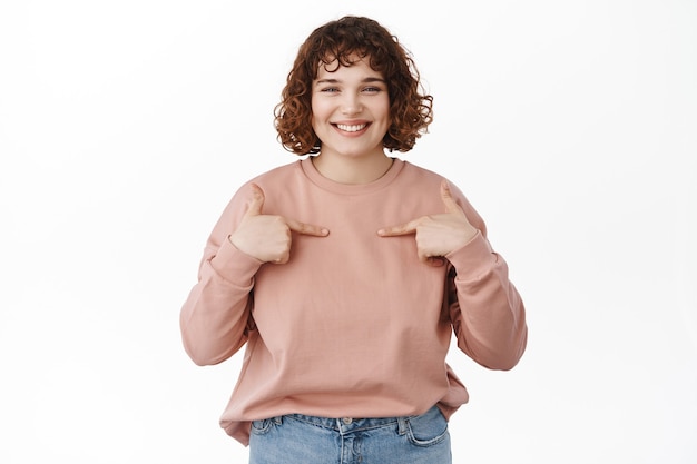 Sonriente niña amable con cabello rizado, apuntando a sí misma con cara feliz, elígeme, soy tu candidato perfecto de pie en blanco