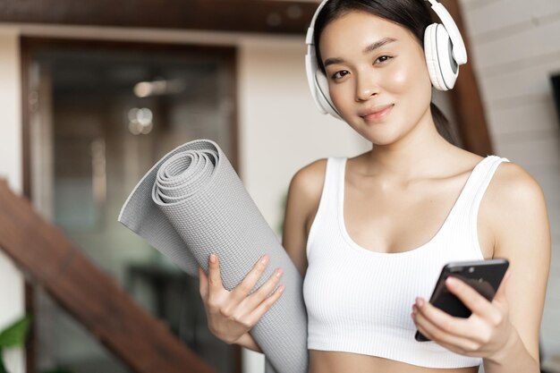 Sonriente niña asiática en auriculares escuchando música en casa y entrenamiento con tapete sosteniendo teléfono móvil ...