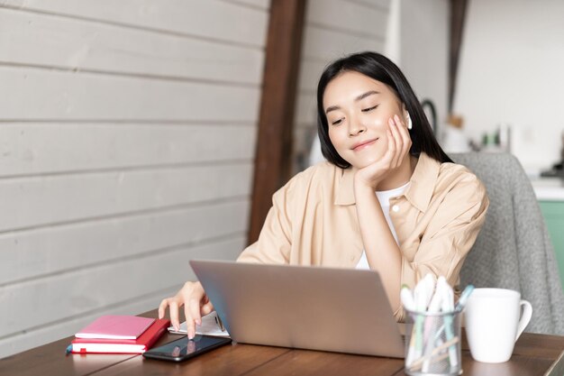 Sonriente niña asiática estudiante mujer sentada con la computadora portátil en casa cocina navegando teléfono inteligente redes sociales ...