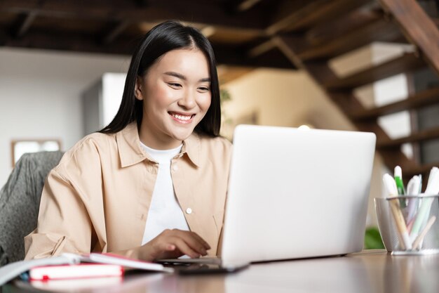 Sonriente niña asiática waching webinar teniendo trabajo videollamada desde casa trabajando freelance remotamente mirando ...