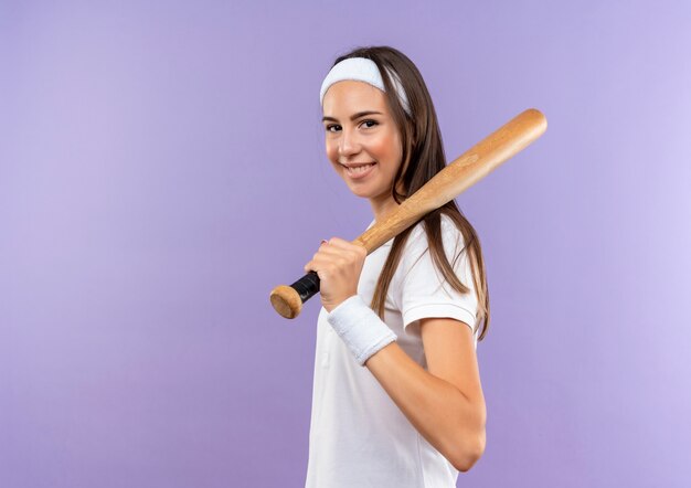 Sonriente niña bastante deportiva con diadema y muñequera sosteniendo un bate de béisbol en el hombro de pie en la vista de perfil aislado en el espacio púrpura