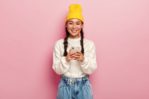 Sonriente niña bonita milenaria usa un teléfono móvil moderno, conectado a internet inalámbrico, descarga imágenes, verifica el buzón de correo electrónico, usa un sombrero amarillo