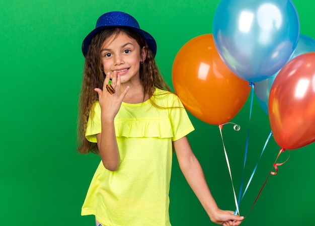 Sonriente niña caucásica con gorro de fiesta azul sosteniendo globos de helio y silbato de fiesta aislado en la pared verde con espacio de copia