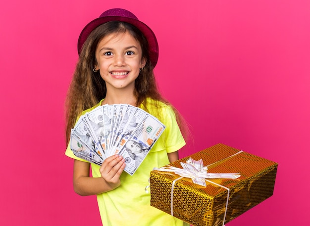 Sonriente niña caucásica con gorro de fiesta púrpura con caja de regalo y dinero aislado en la pared rosa con espacio de copia