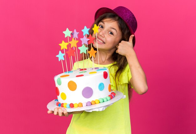 Sonriente niña caucásica con gorro de fiesta púrpura sosteniendo pastel de cumpleaños y pulgar hacia arriba aislado en la pared rosa con espacio de copia