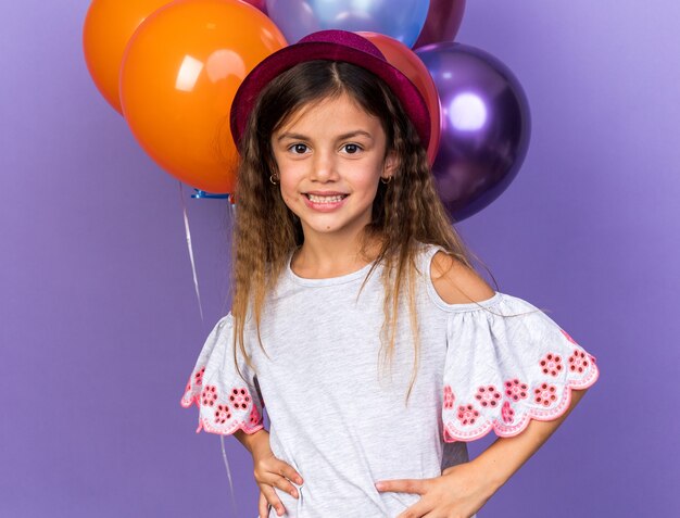 Sonriente niña caucásica con gorro de fiesta violeta de pie delante de globos de helio aislado en la pared púrpura con espacio de copia